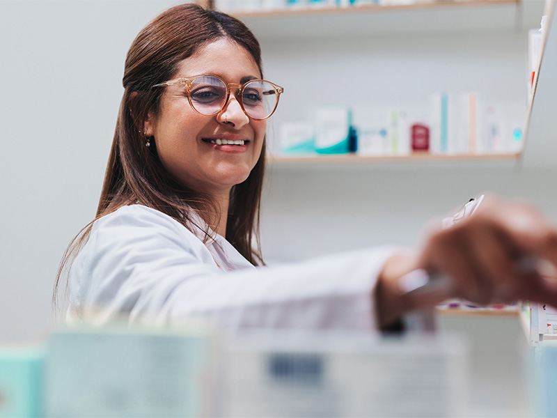 Kvinnlig farmaceut står i ett rum och plockar med läkemedelsförpackningar på en hylla, ser glad ut. 