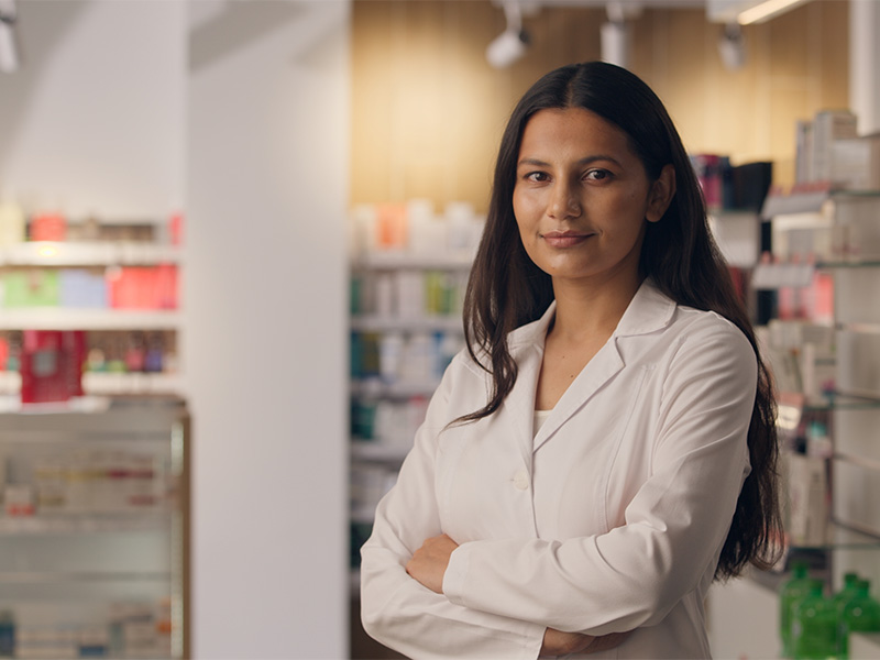 Kvinnlig farmaceut står mitt på ett apotek med dämpat ljus, hon ser belåten och lugn ut.