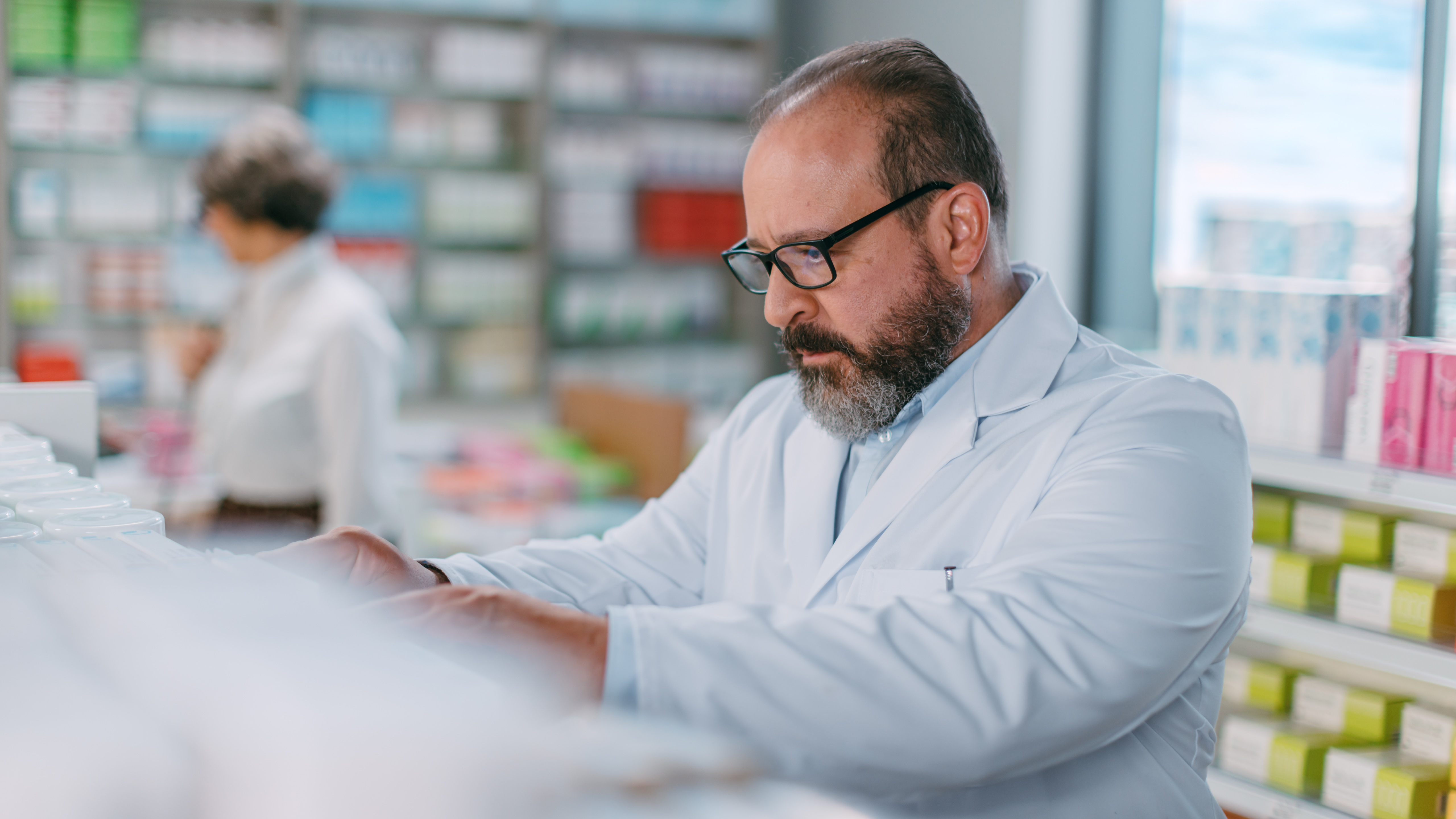 Manlig farmaceut på apotek, plockar med varor på en hylla ute på apoteksgolvet. Ser koncentrerad ut.