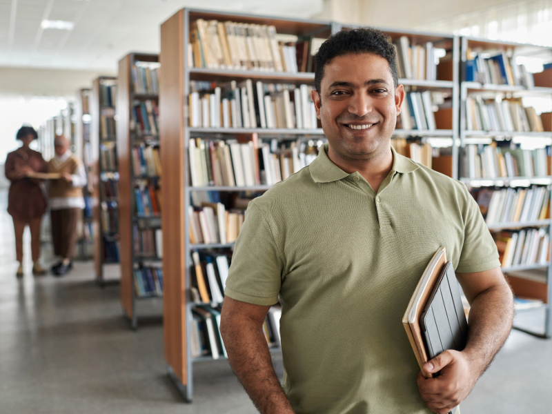 Bild på en man i ett bibliotek som håller i böcker och en ipad, ler och ser nöjd ut. 